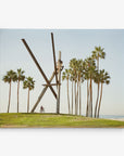 A large, abstract metal sculpture sits on a grassy hill near a beach, surrounded by tall palm trees. A person is riding a bicycle in front of the sculpture. The sky is clear and the ocean is visible in the background, creating a scene perfect for Offley Green's 11x14 California Canvas Print (Choose from 10+ Designs).