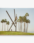 A cyclist rides by a large abstract metal sculpture on a grassy area lined with tall palm trees under a clear sky. The sculpture, reminiscent of 16x20 California Canvas Print (Choose from 10+ Designs) by Offley Green, consists of long intersecting beams with a circular shape near the top. The ocean is slightly visible in the background.