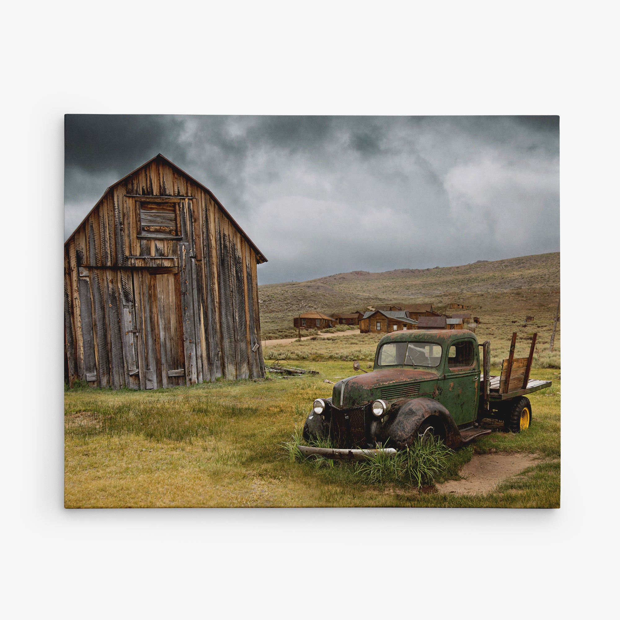 A weathered, vintage truck sits abandoned in a grassy field beside an old, dilapidated wooden barn. In the background, additional rustic wooden structures are visible under a cloudy, overcast sky. This scene evokes a sense of nostalgia and abandonment, perfect for the Offley Green 11x14 Rustic Canvas Print (Choose from 10+ Designs).