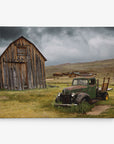 A weathered wooden barn stands on a grassy field under a cloudy sky, captured beautifully as rustic wall art on premium artist-grade canvas. In the foreground, an old, rusted green truck overgrown with weeds is parked, with other aged wooden structures in the background hinting at an abandoned settlement. This scene is rendered exquisitely in the 24x30 Rustic Canvas Print (Choose from 10+ Designs) by Offley Green.