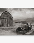 A sepia-toned photograph depicts an old, dilapidated truck partially covered in grass, parked in front of a weathered wooden barn. In the background, rustic buildings dot the cloudy sky. This scene of an abandoned rural area makes for stunning wall art or canvas gallery wrap, perfect for those who appreciate Offley Green's 11x14 Rustic Canvas Print (Choose from 10+ Designs).