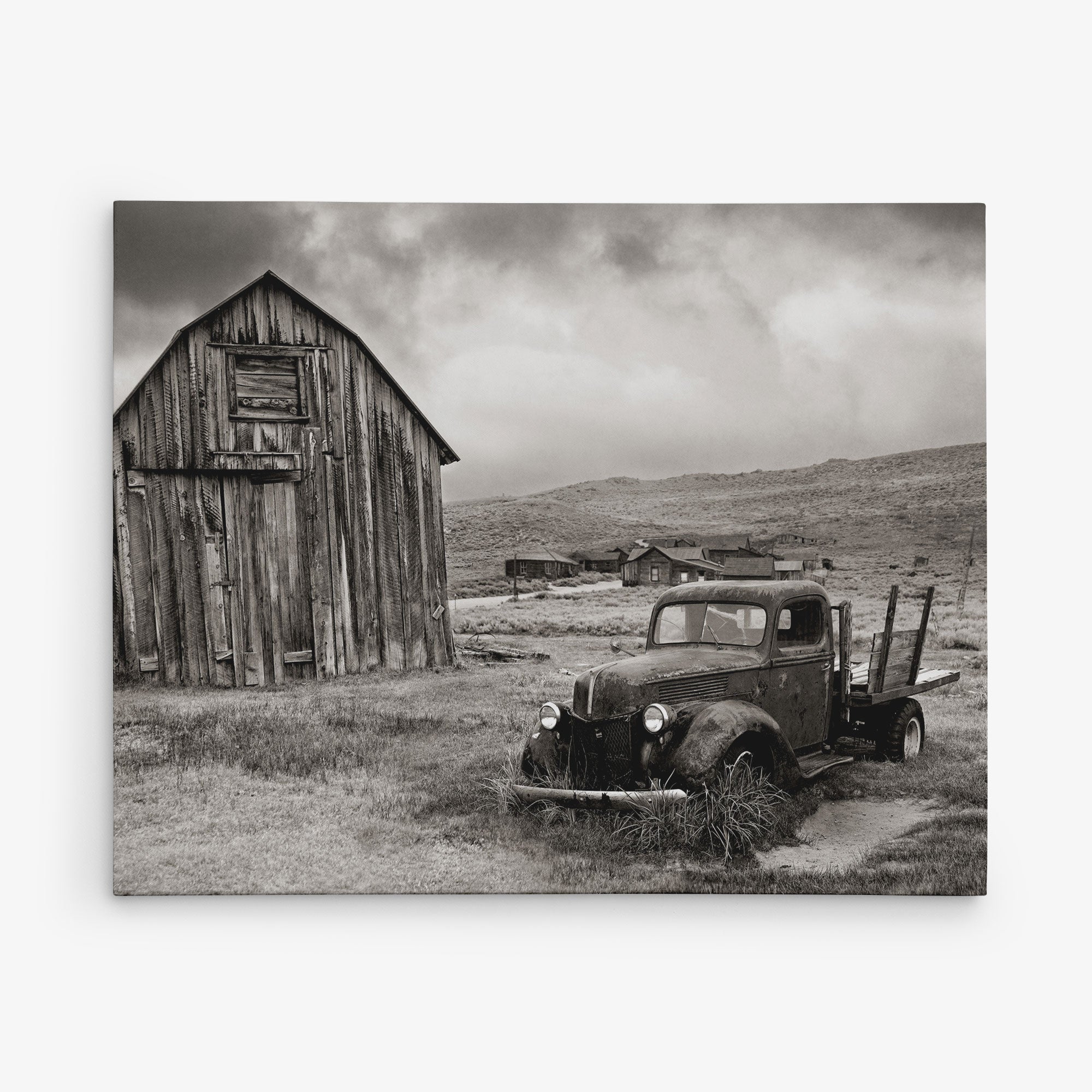 Black and white photograph of an old, rusted truck parked on overgrown grass in front of a weathered wooden barn. In the background, several smaller wooden buildings are scattered across a barren, hilly landscape under a cloudy sky. This piece makes for perfect rustic wall art on premium artist-grade canvas. Introducing the 24x30 Rustic Canvas Print (Choose from 10+ Designs) by Offley Green.