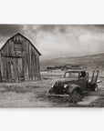 Black and white photograph of an old, rusted truck parked on overgrown grass in front of a weathered wooden barn. In the background, several smaller wooden buildings are scattered across a barren, hilly landscape under a cloudy sky. This piece makes for perfect rustic wall art on premium artist-grade canvas. Introducing the 24x30 Rustic Canvas Print (Choose from 10+ Designs) by Offley Green.