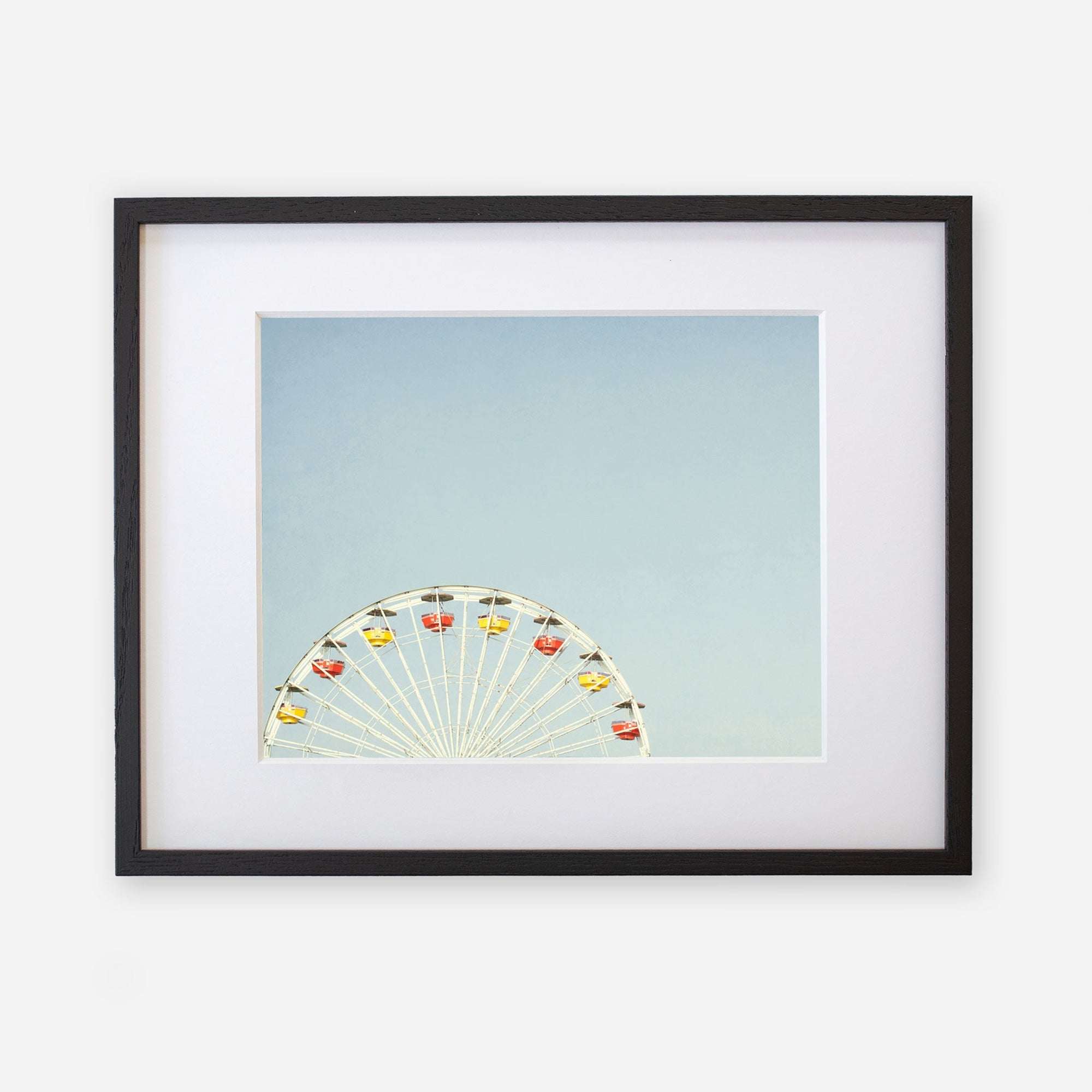 Offley Green&#39;s &#39;Ferris Blue&#39; framed photograph of a ferris wheel against a clear sky at Santa Monica Pier, with the wheel seen from a side angle, displaying colorful passenger cabins. The frame is black with a white mat