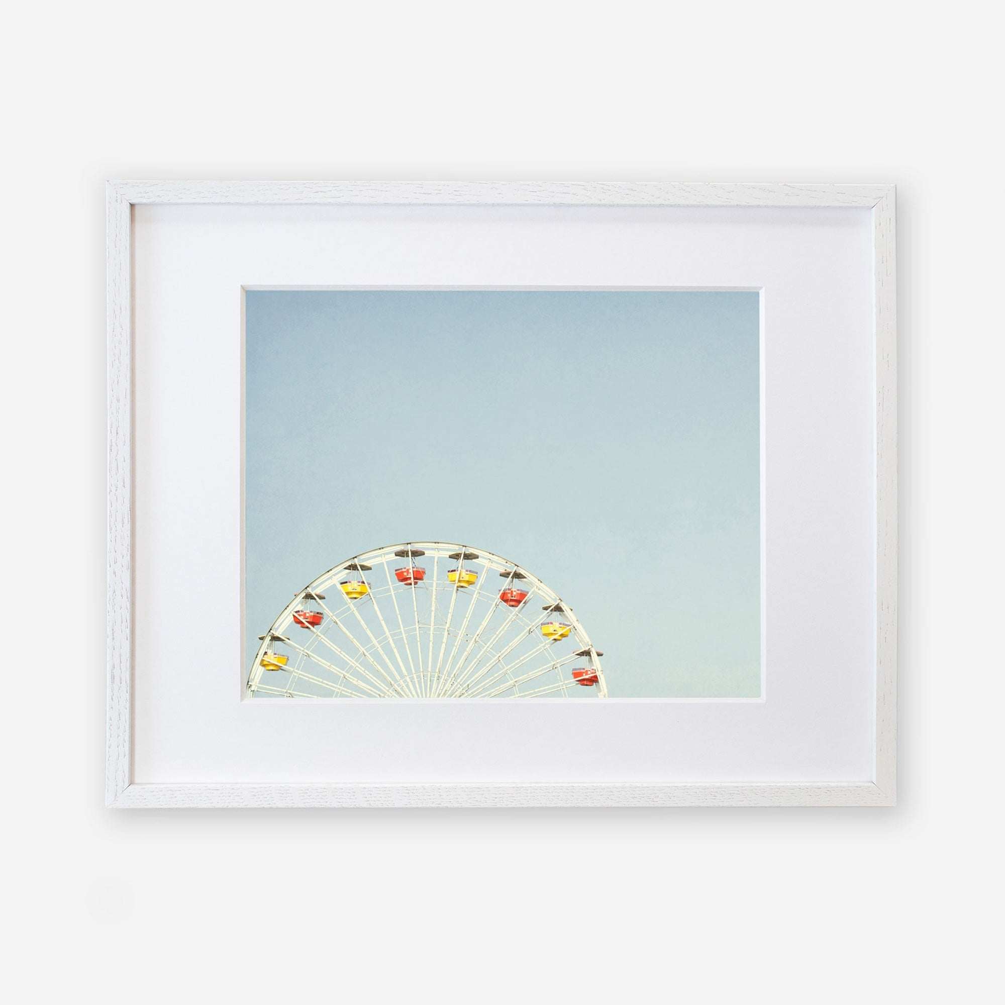 A framed image showcasing the top half of a ferris wheel with colorful gondolas against a pale blue sky, captured on archival photographic paper, centered within a white mat and a simple white frame of Offley Green&#39;s Blue Minimalist Wall Decor, &#39;Ferris Blue&#39;.