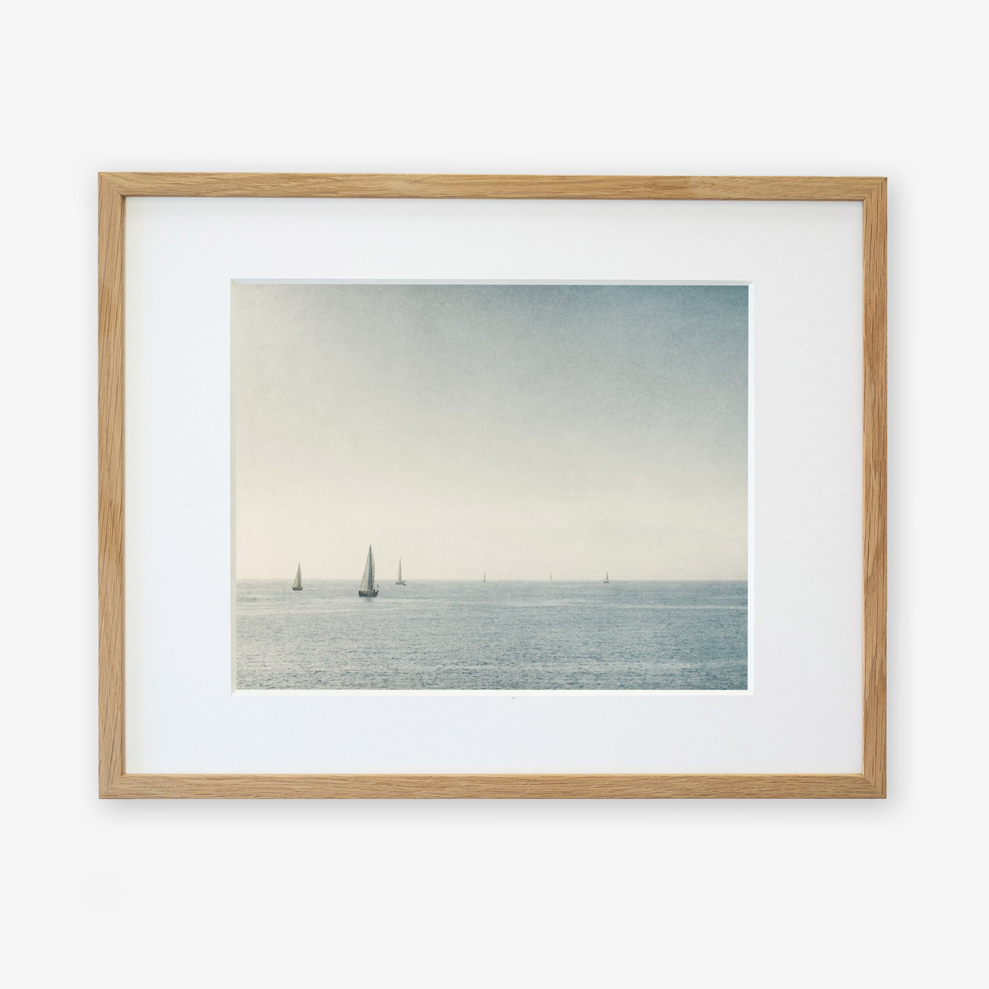 Framed artwork depicting a calm seascape with multiple sailboats on the horizon under a light blue sky, printed on archival photographic paper and enclosed in a light wooden frame with a white mat border: Moody Nautical Seascape Print, 'Sail Boats Approaching' by Offley Green.
