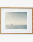 Framed artwork depicting a calm seascape with multiple sailboats on the horizon under a light blue sky, printed on archival photographic paper and enclosed in a light wooden frame with a white mat border: Moody Nautical Seascape Print, 'Sail Boats Approaching' by Offley Green.