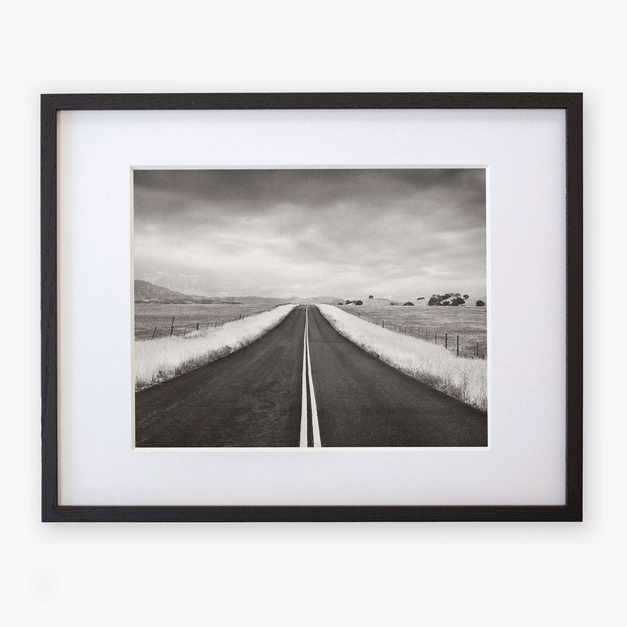 A framed Offley Green black and white photograph of a straight road stretching into the distance through a rural landscape, with grassy fields and scattered trees under a cloudy sky, printed on archival photographic paper.
