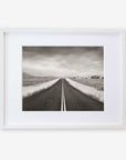 Offley Green's Black and White Rural Landscape Art, 'American Road Trip', featuring a straight road extending to the horizon framed by fields and sparse trees under a cloudy sky, displayed in a white frame on a white background.