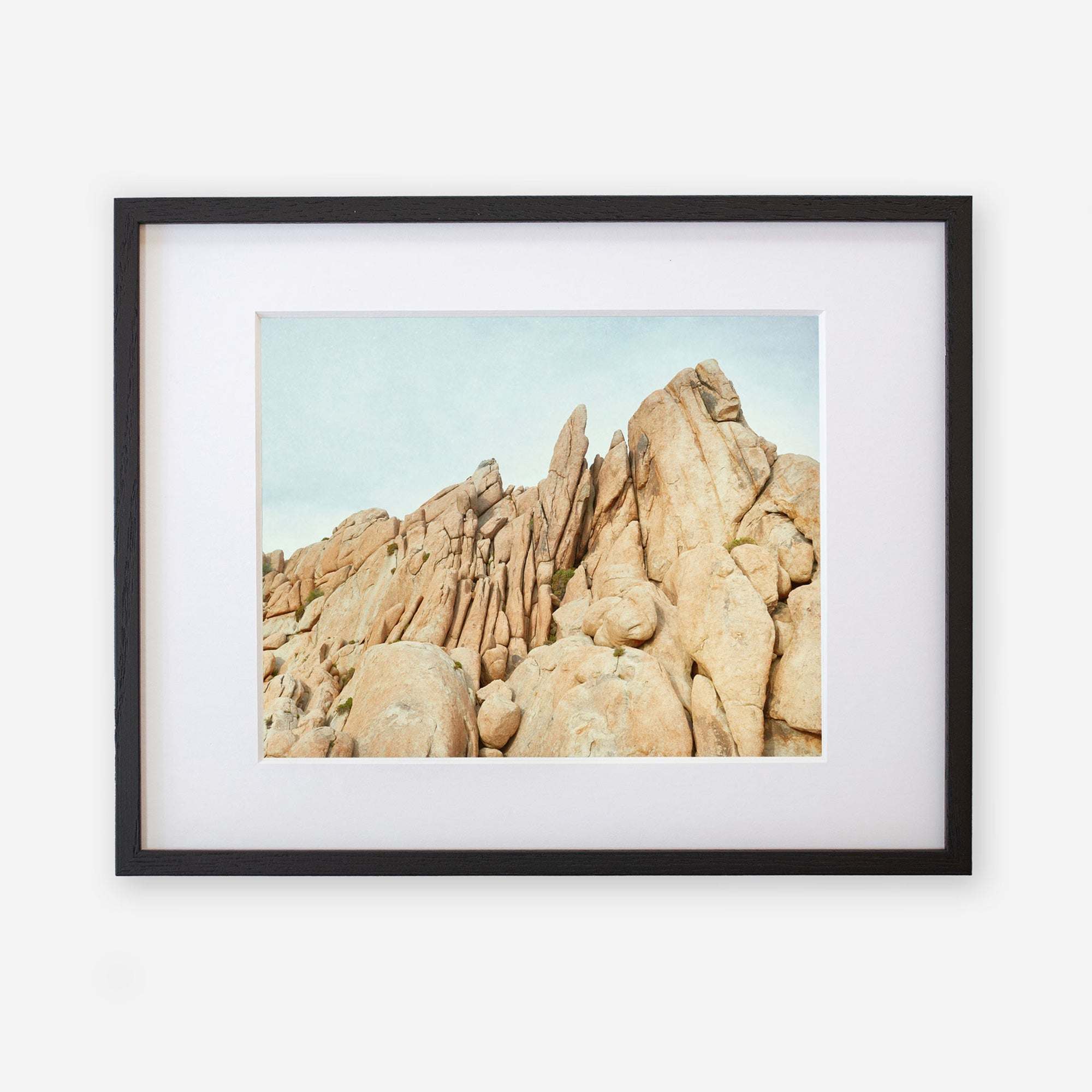 A framed photograph depicting a rugged landscape of towering beige rock formations under a pale blue sky in Joshua Tree. The frame is black with a white mat border, on a white background. - Offley Green&#39;s Joshua Tree Print, &#39;Joshua Rocks&#39;