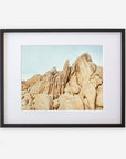 A framed photograph depicting a rugged landscape of towering beige rock formations under a pale blue sky in Joshua Tree. The frame is black with a white mat border, on a white background. - Offley Green's Joshua Tree Print, 'Joshua Rocks'