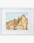 A framed photograph of a rocky desert landscape with towering rock formations under a clear blue sky, printed on archival photographic paper. The frame is white and hangs against a white background. The product is the Offley Green Joshua Tree Print 'Joshua Rocks'.