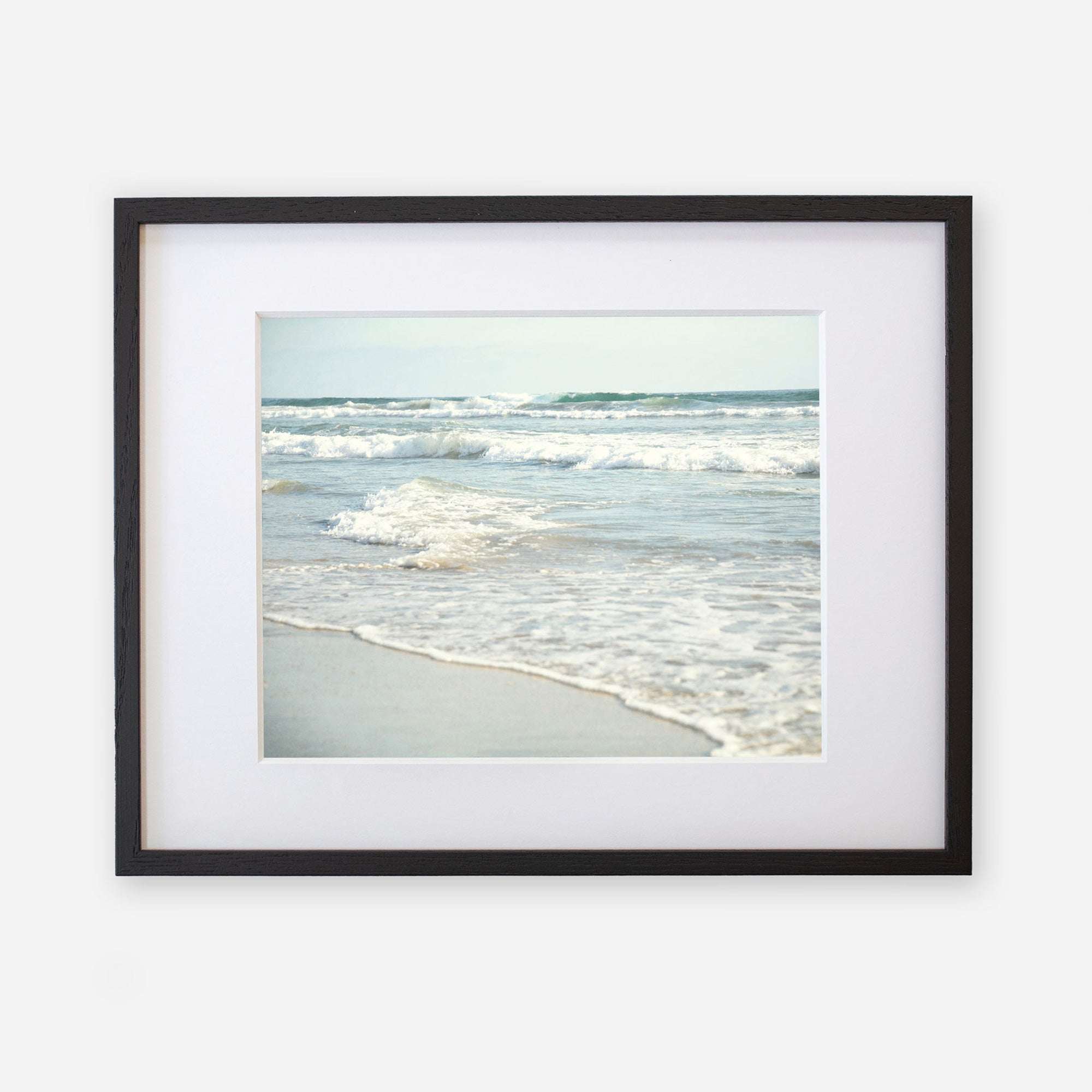 A framed photograph of a serene scene at Carlsbad State Beach, displaying gentle waves breaking near the shore with a clear sky in the background. The frame is black with a white mat featuring the Coastal Beach Print in California &#39;Surf and Sun&#39; by Offley Green.