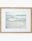 A framed photograph of a tranquil Carlsbad State Beach scene, with gentle waves lapping at the shore under a clear sky. The frame is simple and made of light wood, mounted on a white Offley Green Coastal Beach Print in California 'Surf and Sun'.