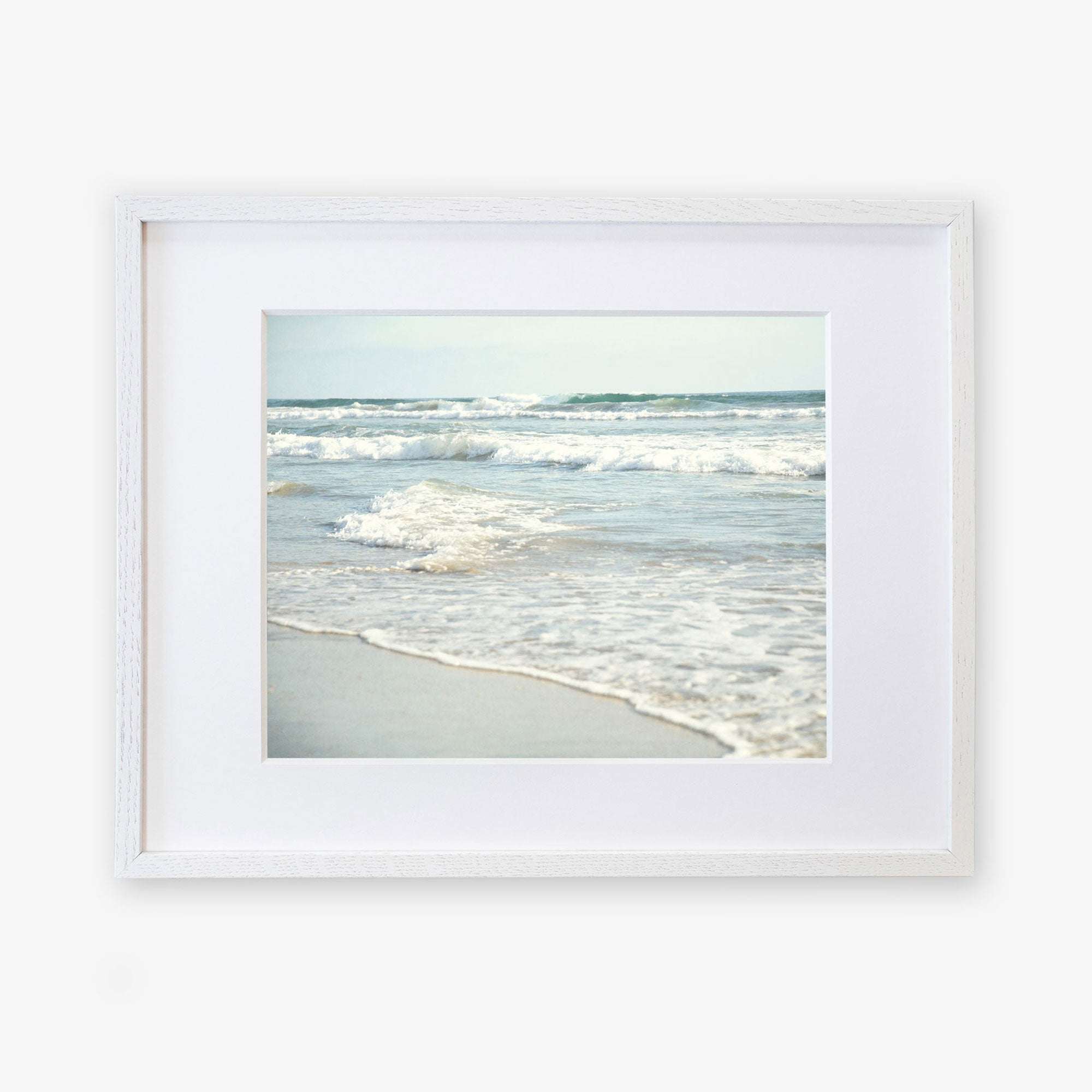 A framed photograph of Carlsbad State Beach with gentle waves and soft sand, displayed against a white background. The frame is simple and white, enhancing the tranquil Coastal Beach Print in California &#39;Surf and Sun&#39; by Offley Green.