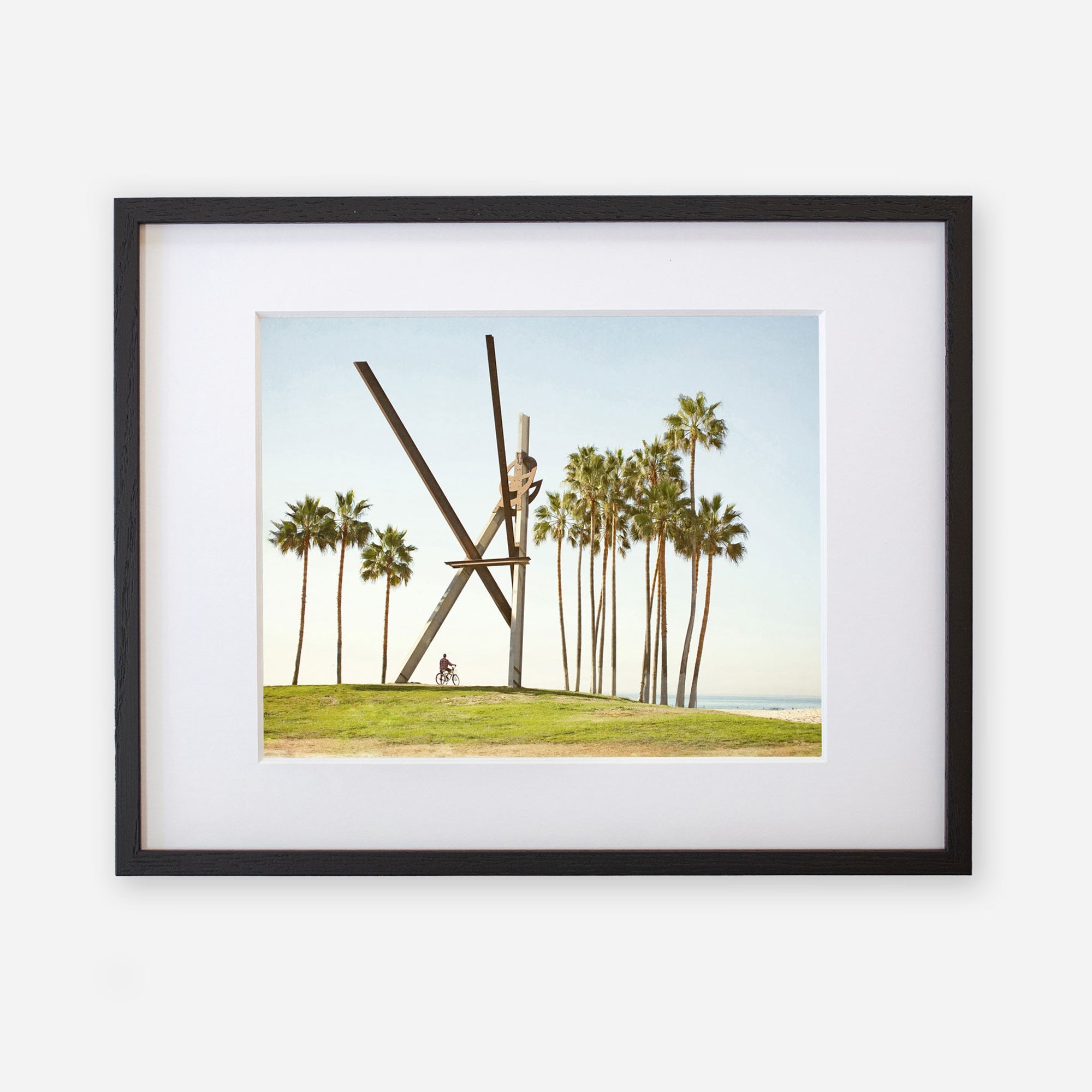 Unframed photograph of an outdoor scene featuring tall palm trees and a large modern sculpture resembling an &quot;x&quot; shape, set against a clear sky, with a small figure seated near the base of the Offley Green Venice Beach Landmark Sculpture, &#39;V is for Venice&#39;.
