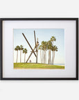 Unframed photograph of an outdoor scene featuring tall palm trees and a large modern sculpture resembling an "x" shape, set against a clear sky, with a small figure seated near the base of the Offley Green Venice Beach Landmark Sculpture, 'V is for Venice'.