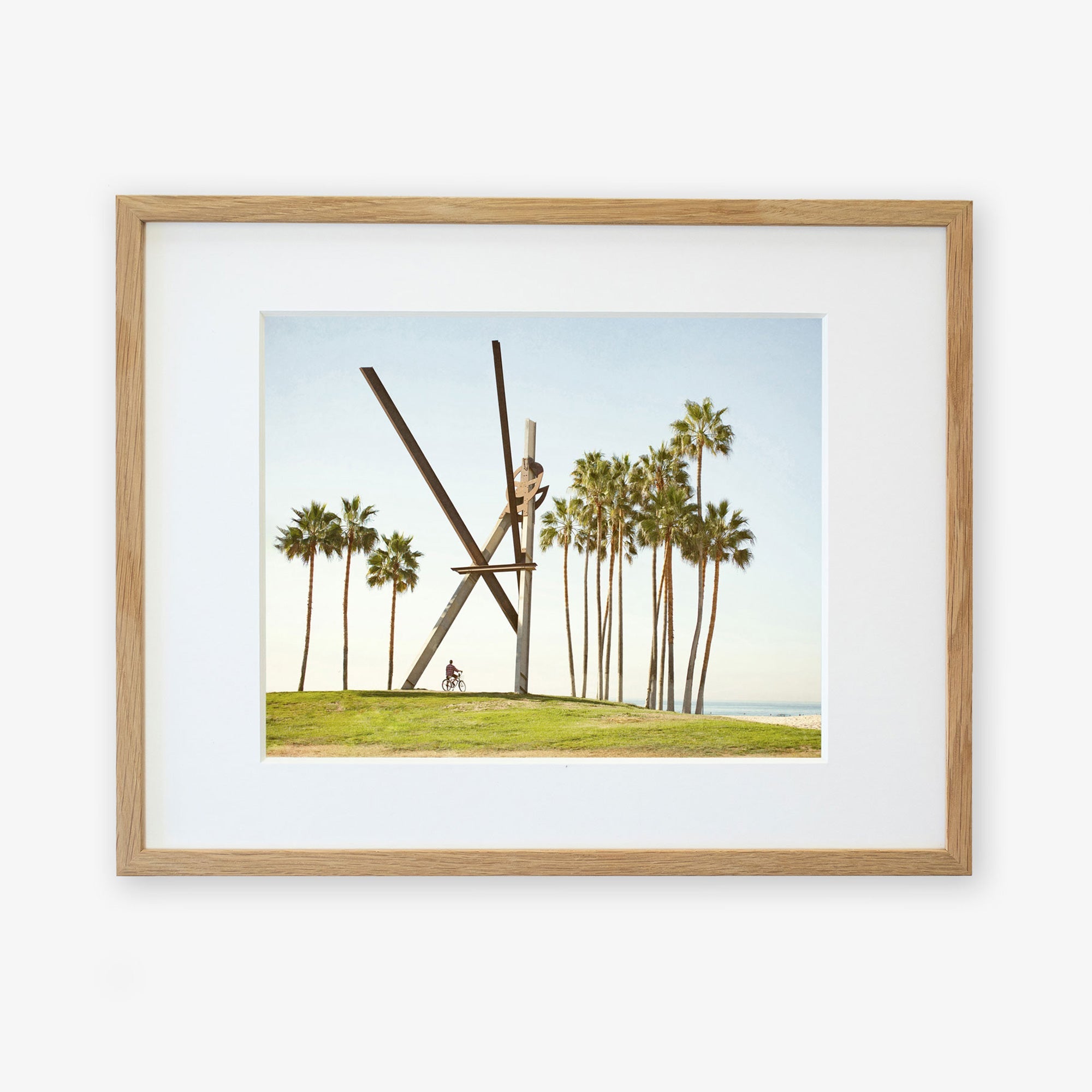 An unframed artwork depicting the Offley Green &#39;V is for Venice&#39; sculpture, a surreal scene with oversized wooden clothespins partially buried in a grassy field, surrounded by tall palm trees under a clear blue sky.