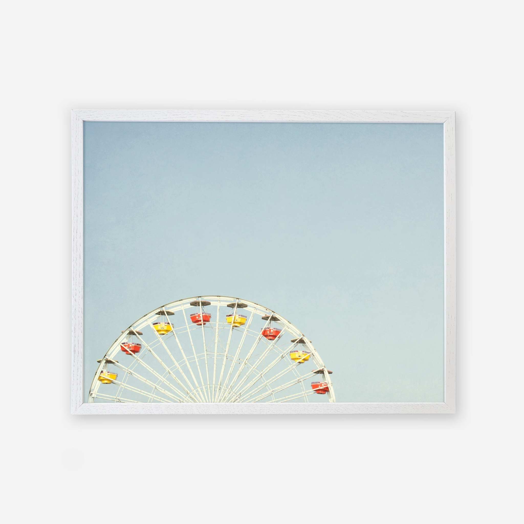 A framed picture of the Blue Minimalist Wall Decor &#39;Ferris Blue&#39; by Offley Green, with a white ferris wheel and yellow and red gondolas at Santa Monica Pier, half-visible against a clear blue sky.