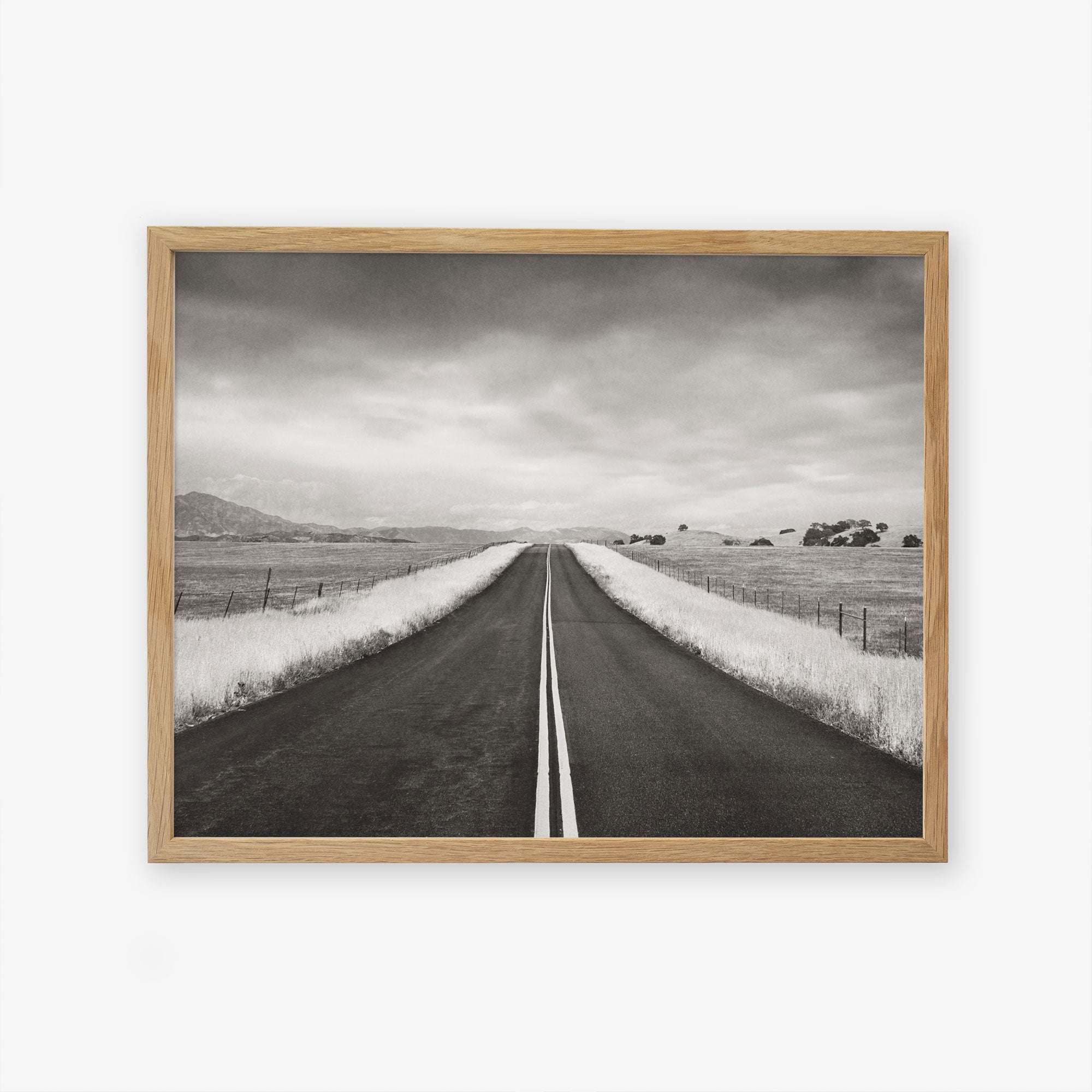 A framed Offley Green black and white photograph on archival photographic paper of a straight road leading through a rural landscape with fenced fields and sparse trees under a cloudy sky, titled &#39;American Road Trip&#39;.