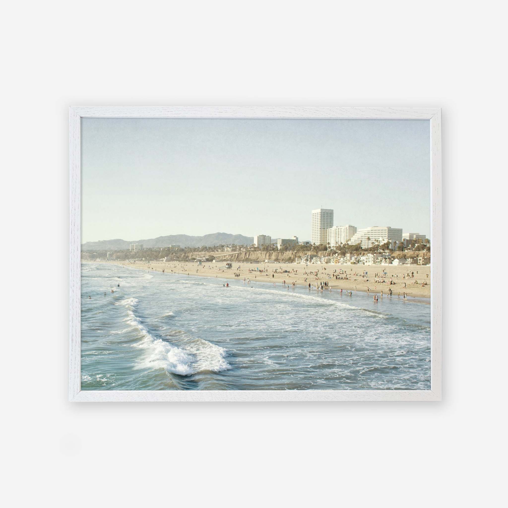 A framed photograph of a busy beach scene with people scattered along the shore and in the water, waves gently breaking, and a city skyline in the background under a clear sky, printed on archival photographic paper. Offley Green&#39;s Santa Monica Print, &#39;Santa Monica Seaside&#39;.