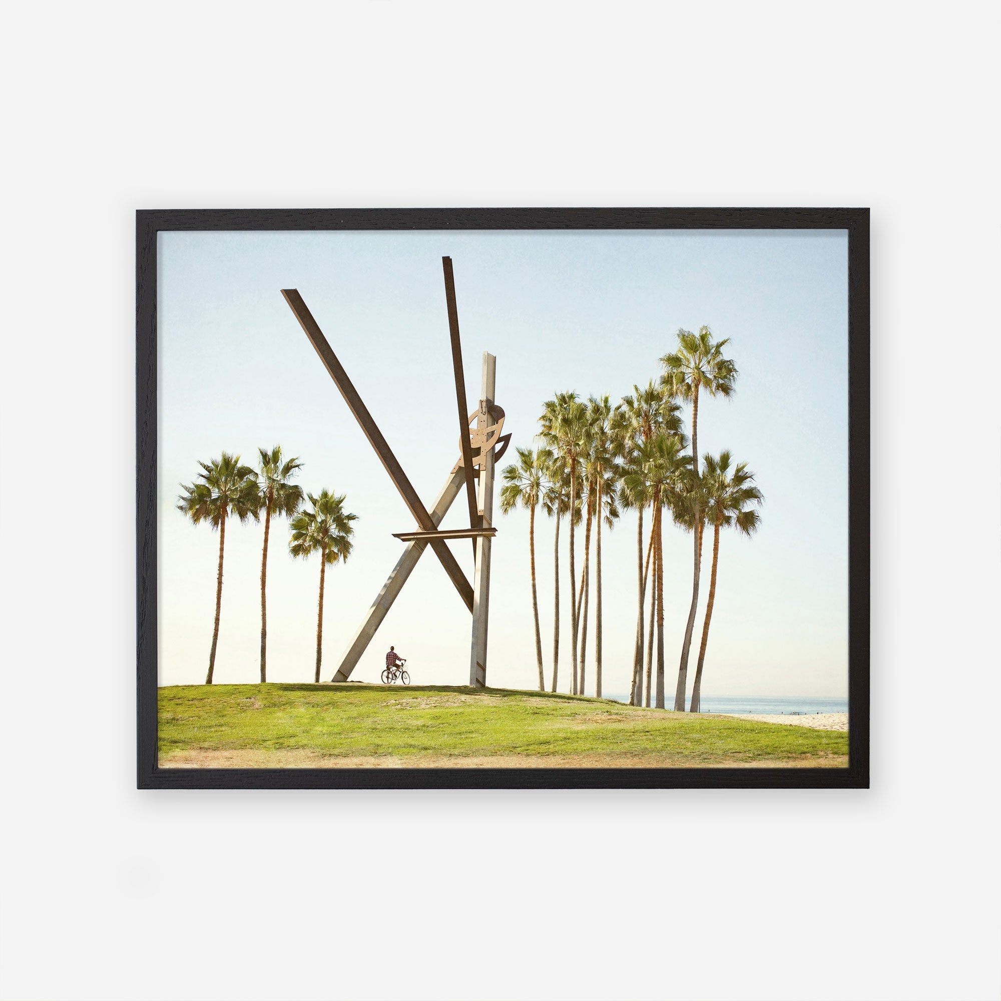 A framed artwork depicting the Venice Beach Landmark Sculpture, &#39;V is for Venice&#39;, by Offley Green on a grassy knoll near Venice Beach, surrounded by tall palm trees, with a clear sky in the background and a cyclist at its base.