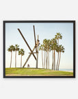 A framed artwork depicting the Venice Beach Landmark Sculpture, 'V is for Venice', by Offley Green on a grassy knoll near Venice Beach, surrounded by tall palm trees, with a clear sky in the background and a cyclist at its base.