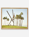 A framed artistic depiction of the Venice Beach Landmark Sculpture, 'V is for Venice', by Offley Green, with intersecting beams, positioned on Venice Beach with palm trees and a clear sky in the background, and a cyclist in the foreground.