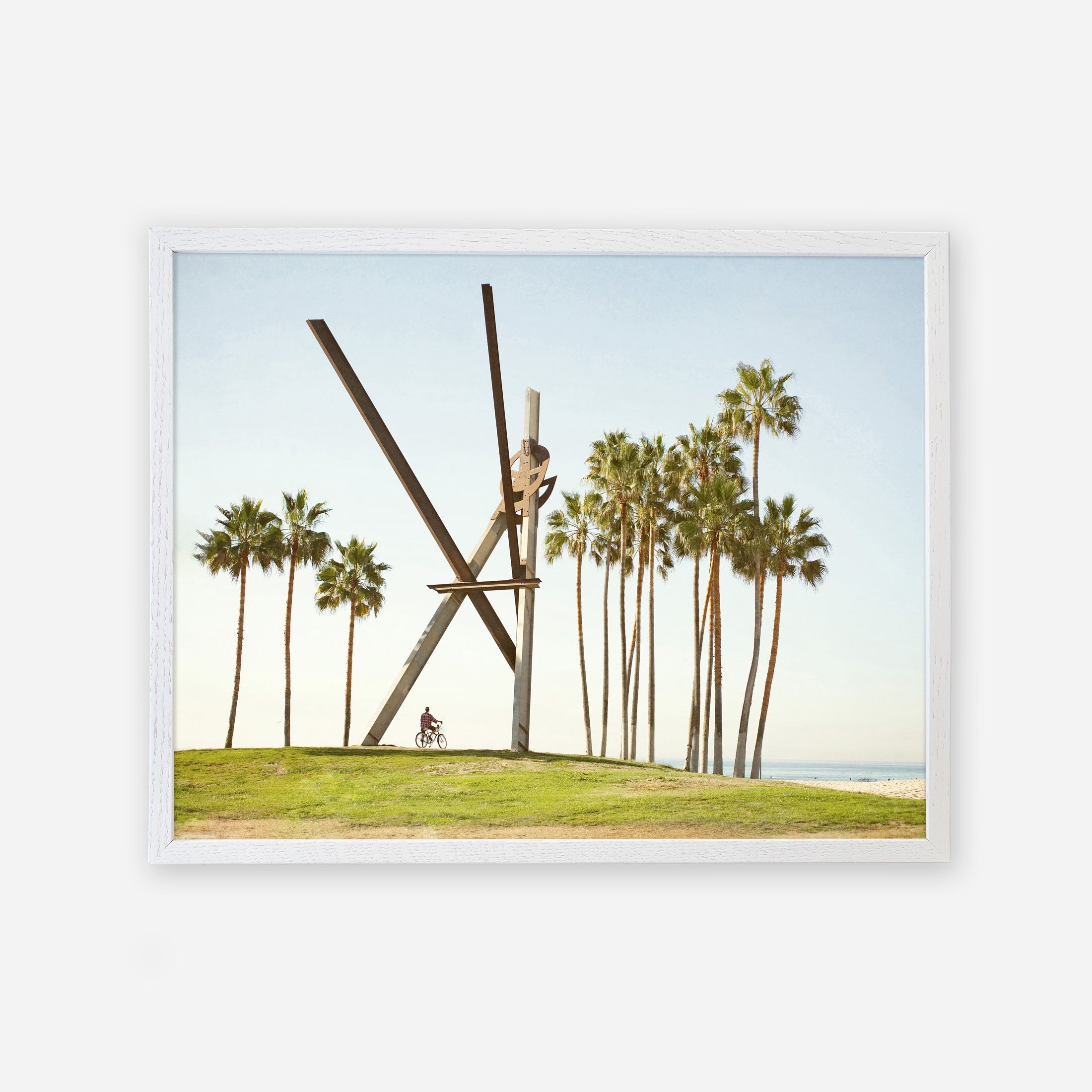 A framed photograph of the Venice Beach Landmark Sculpture, &#39;V is for Venice&#39; by Offley Green, resembling intersecting wooden beams, with tall palm trees in the background under a clear sky. This Venice Beach photography captures a cyclist riding by on a pathway.