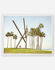 A framed photograph of the Venice Beach Landmark Sculpture, 'V is for Venice' by Offley Green, resembling intersecting wooden beams, with tall palm trees in the background under a clear sky. This Venice Beach photography captures a cyclist riding by on a pathway.