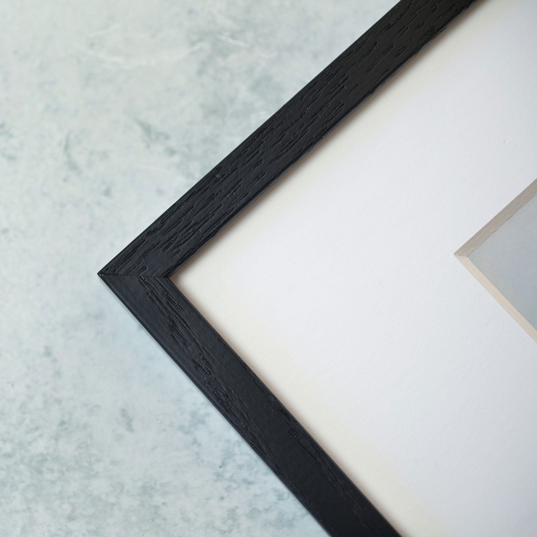 A close-up view of a black wooden picture frame corner against a light, marbled background. The frame, housing an Offley Green product named Rustic Floral Print, &#39;Fields of Lavender&#39; on archival photographic paper, features white matting inside that&#39;s partially visible.