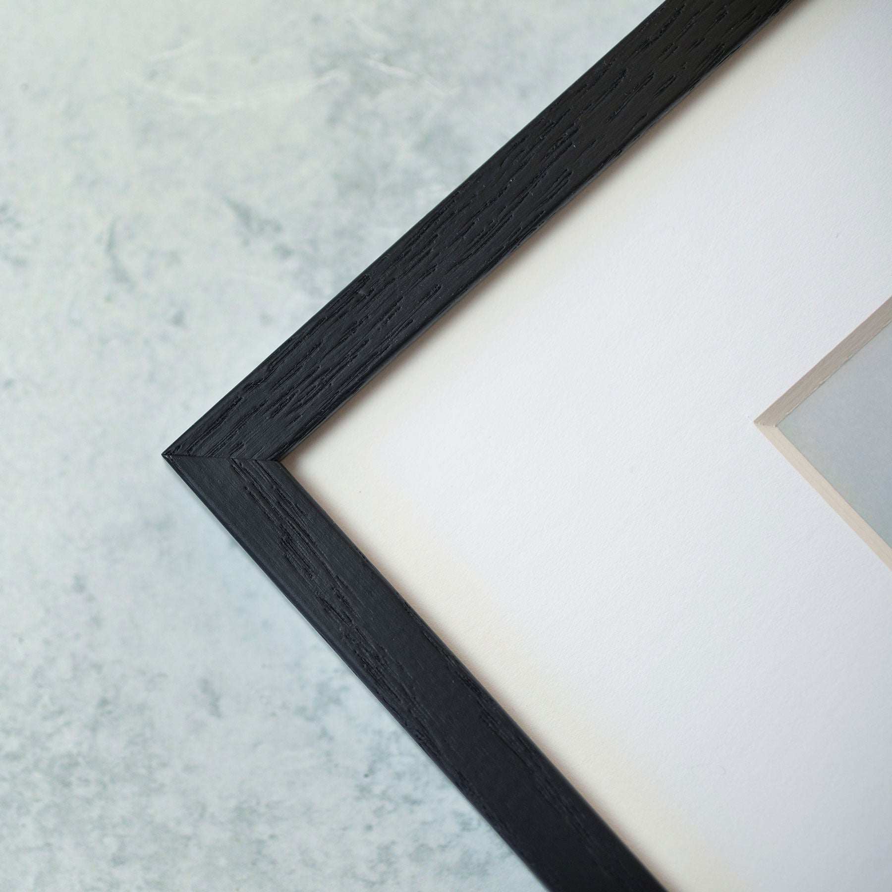 A close-up view of the corner of a picture frame. The frame, made of black wood with visible grain texture, surrounds a white matte border containing the Rustic Countryside Print, 'Tree in a Field' by Offley Green. The background is a textured light gray surface reminiscent of storm clouds.