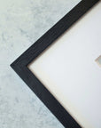Close-up of a corner of a black textured Offley Green picture frame on a textured light gray background, displaying a part of white canvas and a small section of archival photographic paper featuring the California Venice Beach Print, 'Three Palms'.