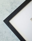 Close-up of a black textured Offley Green picture frame on a subtly textured pale blue background, focusing on the top left corner where the frame meets blank archival photographic paper.