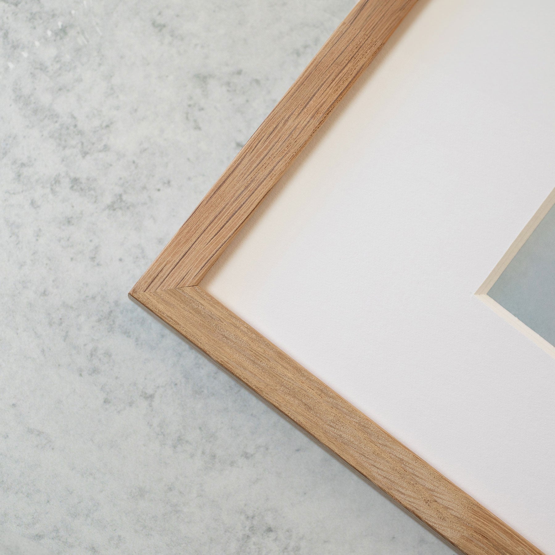 Close-up of a wooden picture frame corner resting on a light gray speckled surface. The frame has a clean, simple design and a natural wood finish. Inside the frame, Offley Green&#39;s &#39;Fields of Lavender&#39; Rustic Floral Print is partially visible, featuring a light-colored background.