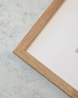 Close-up of a corner of a light wooden picture frame with a white mat bordering the image inside. The frame, set on a white marble surface, showcases the wood's grain and the mat's clean lines, creating a serene atmosphere. The photo is printed on archival photographic paper for timeless quality and features the Nautical Sail Boat Art, 'Sailing Into Rain' by Offley Green.
