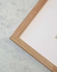 Close-up of the corner of a light wooden picture frame with a white matting, sitting on a grey, marbled surface. The image inside the frame is partially visible on the right edge, printed on archival photographic paper: Rustic Countryside Print, 'Tree in a Field' by Offley Green.