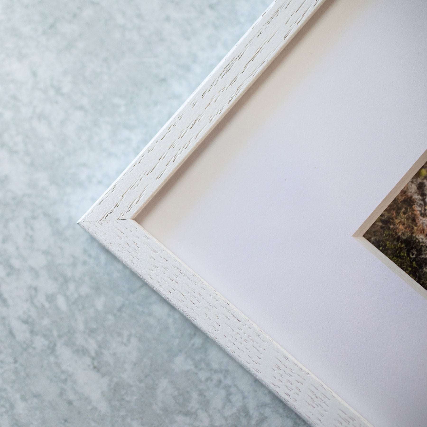 Close-up photo of the corner of a white picture frame set against a light gray, textured background. The frame has a simple, clean design, and the image within shows the edge of an Offley Green Rustic Countryside Print, &#39;Tree in a Field&#39; on archival photographic paper with part of an indeterminate natural scene.