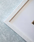 Close-up of a white textured picture frame corner on a gray background, partially showing an Offley Green 'Joshua Rocks' photography print with natural elements.