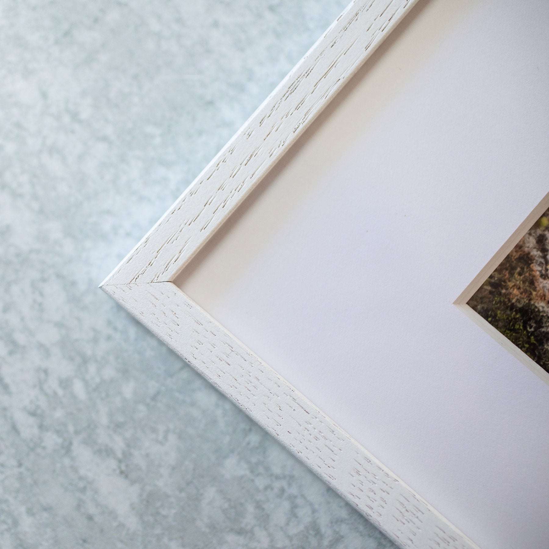 A close-up shot of the corner of a white picture frame. The frame has a textured finish, and part of the enclosed Moody Nautical Seascape Print, &#39;Sail Boats Approaching&#39; by Offley Green is visible on the right side. The background is a light, speckled surface.