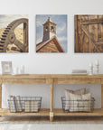 A wooden console table against a white wall displays Offley Green's Set of 3 Canvas featuring a gear, clock tower, and wooden door. The table also holds a framed quote, candles, white vases, and two wire baskets with pillows. Two closed doors flank the scene.