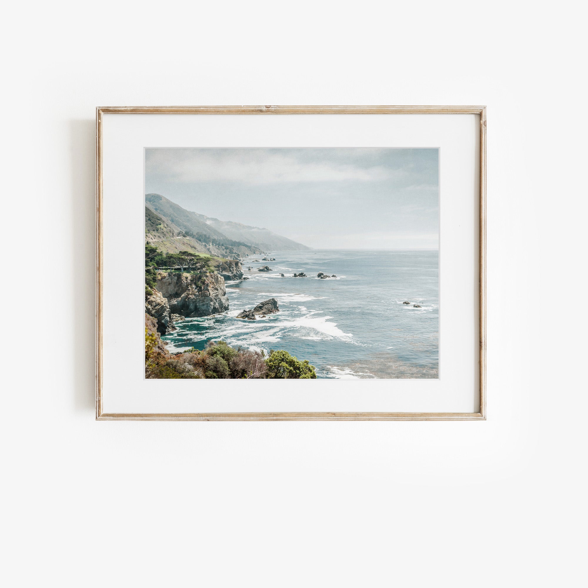 A framed landscape photograph from Offley Green's Big Sur Landscape Print collection, titled 'Rocky Rocks,' hangs on a white wall. Printed on archival photographic paper, the image depicts a coastal scene with rocky cliffs, green foliage, and an ocean where waves crash against the shore. Hazy, overcast skies complete the serene setting reminiscent of California's Highway 1.