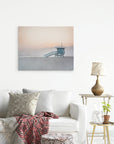 A cozy living room featuring a white sofa with patterned throw pillows and a red patterned blanket. A small side table with a lamp, a potted plant, and decor items sits beside the sofa. A large 24x30 California Canvas Print (Choose from 10+ Designs) by Offley Green of a lifeguard tower by the ocean hangs on the wall, adding to the serene ambiance.