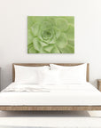 A minimalist bedroom features a wooden bed with white linens, flanked by two wooden nightstands. Each nightstand has a lamp and a small decorative item. Mounted above the bed is a stunning 30X40 Botanical Canvas Print (Choose from 10+ Designs) by Offley Green. The floor is white and carpeted.