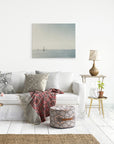 A cozy living room with a white couch adorned with patterned cushions and a red and black throw. A round ottoman with geometric patterns sits on the floor. The wall features a 24x30 Coastal Canvas Print by Offley Green, showcasing one of their minimalist seascape designs. A side table holds a lamp, vase, and small plant.