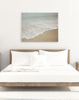 Minimalist bedroom with a wooden bed frame and white bedding. A nightstand on each side of the bed features a small decor item and lamp. A large 30x40 Coastal Canvas Print by Offley Green hangs as wall art above the bed, and a vase with flowers adorns the right nightstand.