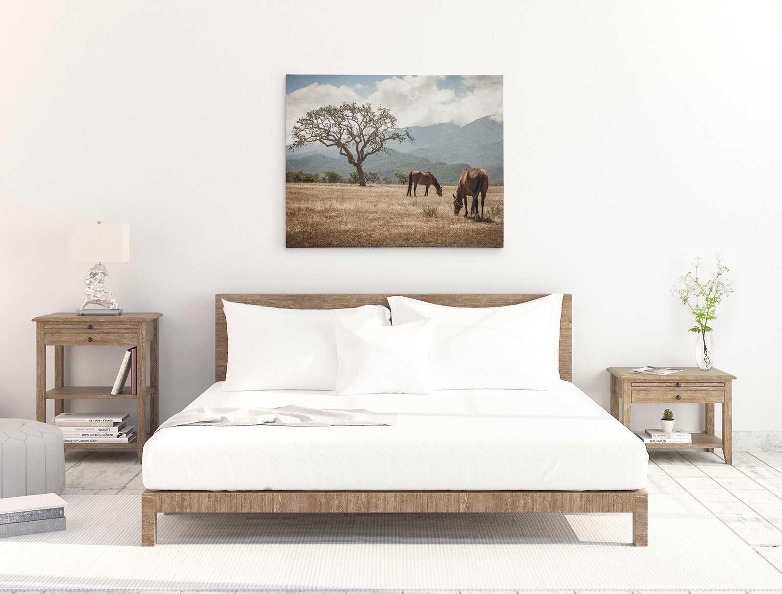 A minimalist bedroom features a wooden bed with white bedding. Flanking the bed are two wooden bedside tables, one with a lamp and the other with a small plant. A ready-to-hang Offley Green 30x40 Rustic Canvas Print (Choose from 10+ Designs) of horses grazing in a field hangs on the wall above the bed.