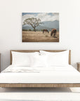 A minimalist bedroom features a wooden bed with white bedding. Flanking the bed are two wooden bedside tables, one with a lamp and the other with a small plant. A ready-to-hang Offley Green 30x40 Rustic Canvas Print (Choose from 10+ Designs) of horses grazing in a field hangs on the wall above the bed.