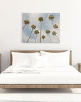 A minimalist bedroom features a wooden bed with white bedding, flanked by two wooden nightstands each holding a lamp. Centering the room is an Offley Green 30x40 California Canvas Print, depicting tall palm trees against a clear sky. Books and decor items adorn the space, creating a cozy atmosphere.