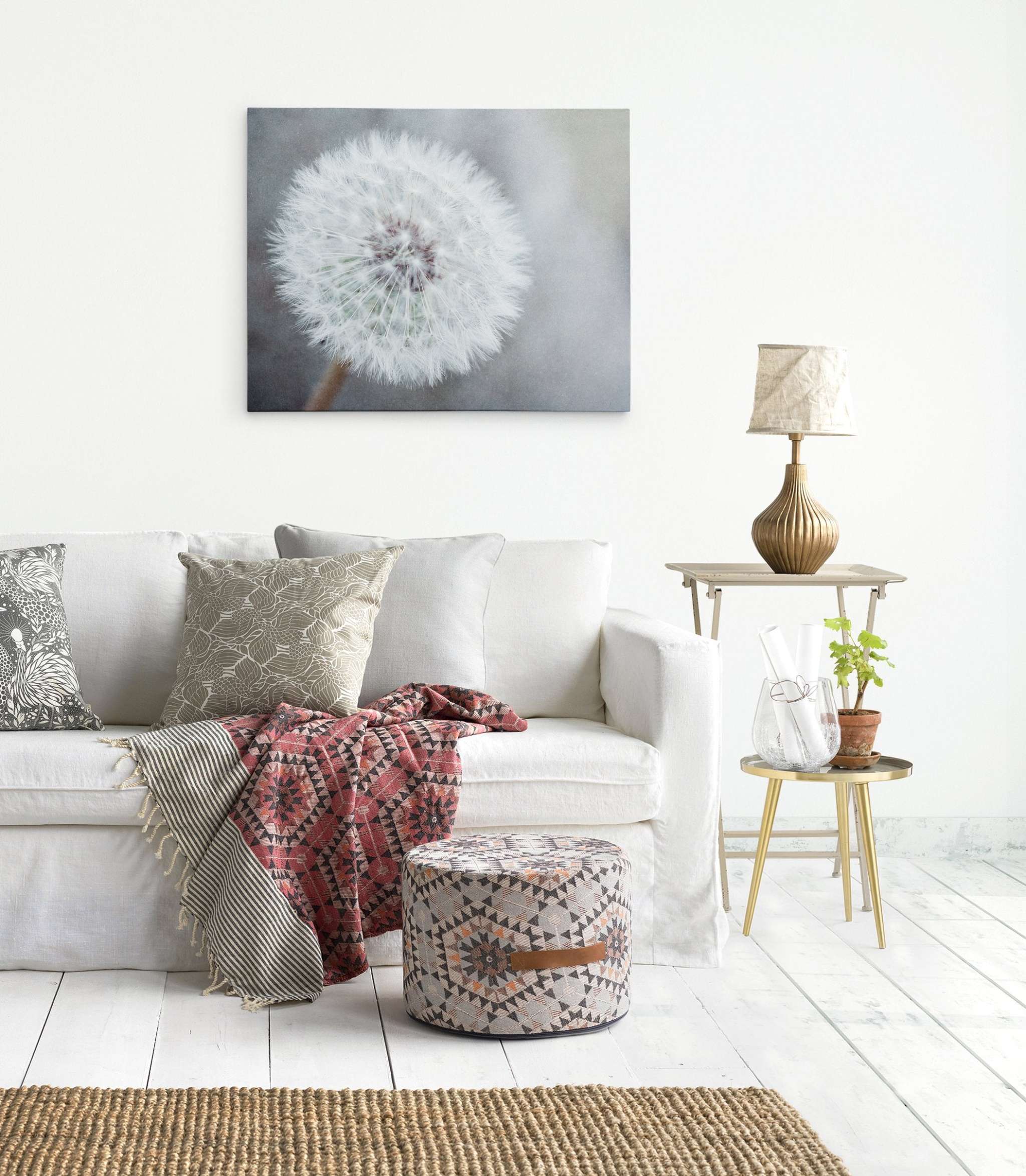 A bright, minimalist living room featuring a white sofa adorned with patterned cushions and a colorful throw blanket. Above the sofa, an Offley Green Neutral Grey Floral Canvas Wall Art, 'Dandelion King' graces the wall. An ornate lamp on a side table and a small plant on a round table beside the sofa complete this ready-to-hang solution.